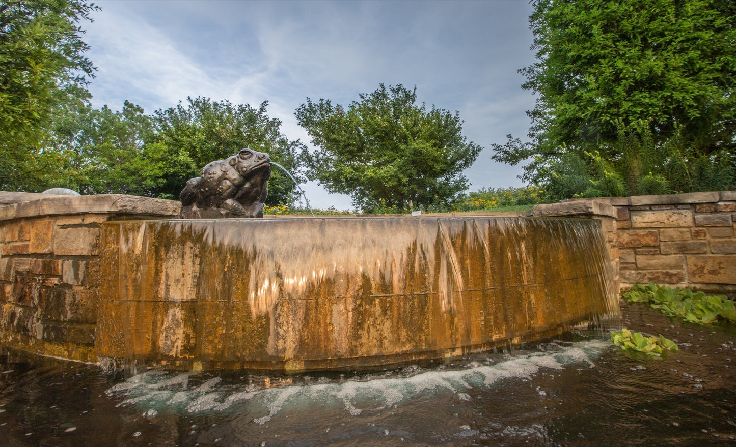 Bronze frog sculpture spouting water on curved stone fountain wall at Powell Gardens botanical garden near Kansas City Missouri