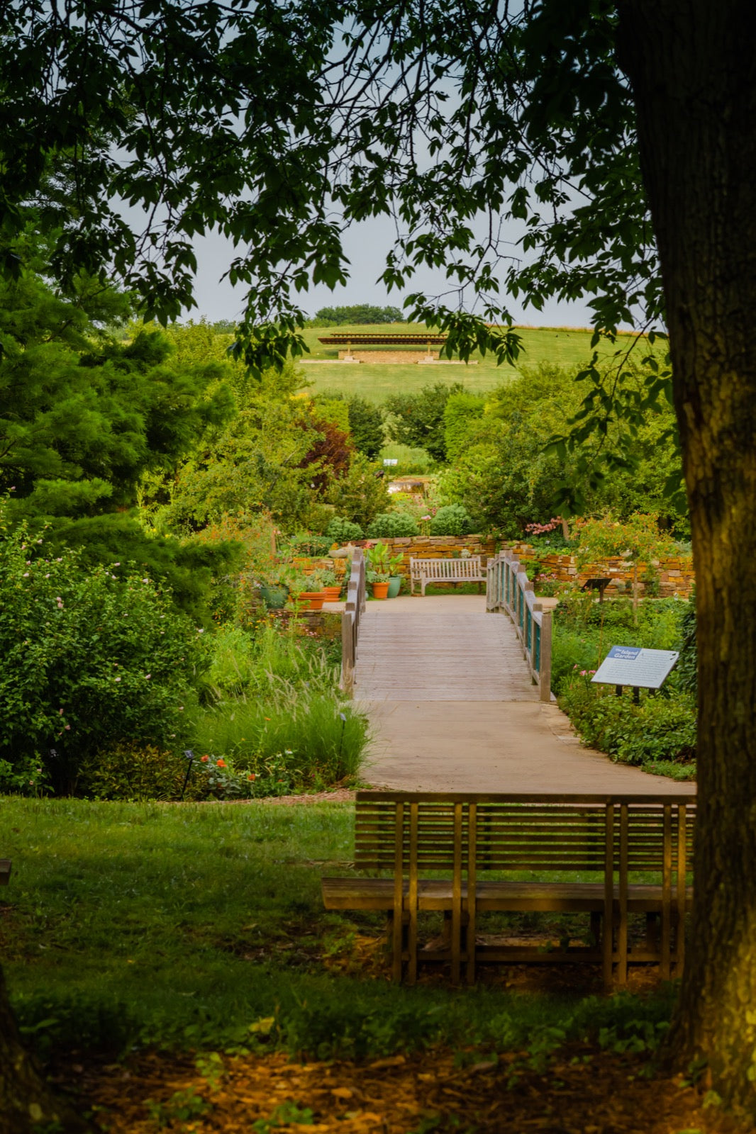 Wooden footbridge path through lush green gardens with potted plants and flower beds at Powell Gardens near Kansas City Missouri