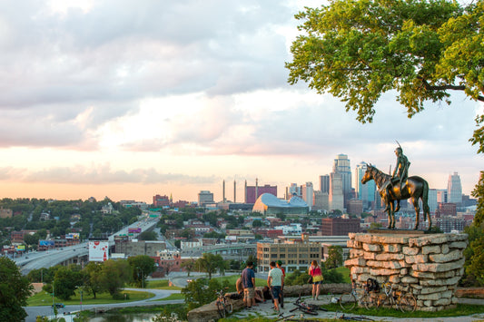 Wide panoramic view of The Scout statue with visitors overlooking the full Kansas City Missouri downtown skyline at sunset from Penn Valley Park